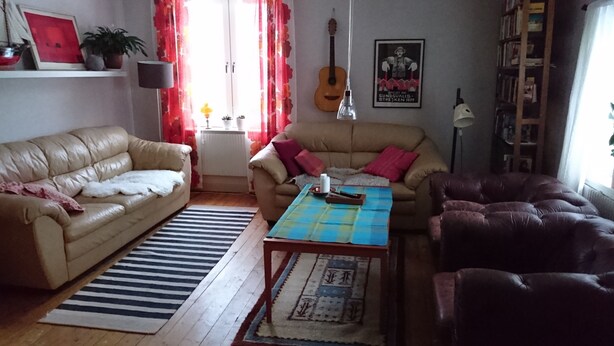 Een woonkamer met een beige bank, een bruine leren fauteuil, een salontafel met een geruit tafelkleed, een gitaar aan de muur en een boekenkast.