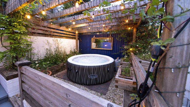 Un jacuzzi en un jardín con terraza de madera, un pérgola de madera con luces colgantes y un espejo en la pared.