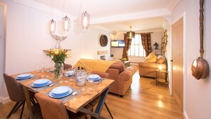 Un comedor con una mesa de madera dispuesta para cuatro personas, rodeada de sillas tapizadas en tono marrón. En el fondo, se ve un televisor montado en la pared, y un sofá es visible en la sala de estar contigua.