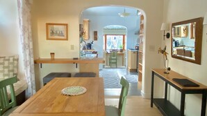 Un coin repas avec une table en bois, des chaises et un lustre. On aperçoit une cuisine à travers une arche en arrière-plan.