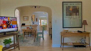 Un salon avec un téléviseur diffusant un journal télévisé, un coin repas avec une table et des chaises, et une affiche encadrée accrochée au mur.