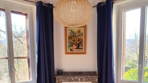 Une pièce avec une cheminée, de grandes fenêtres avec des rideaux, un luminaire suspendu et un tableau au mur.