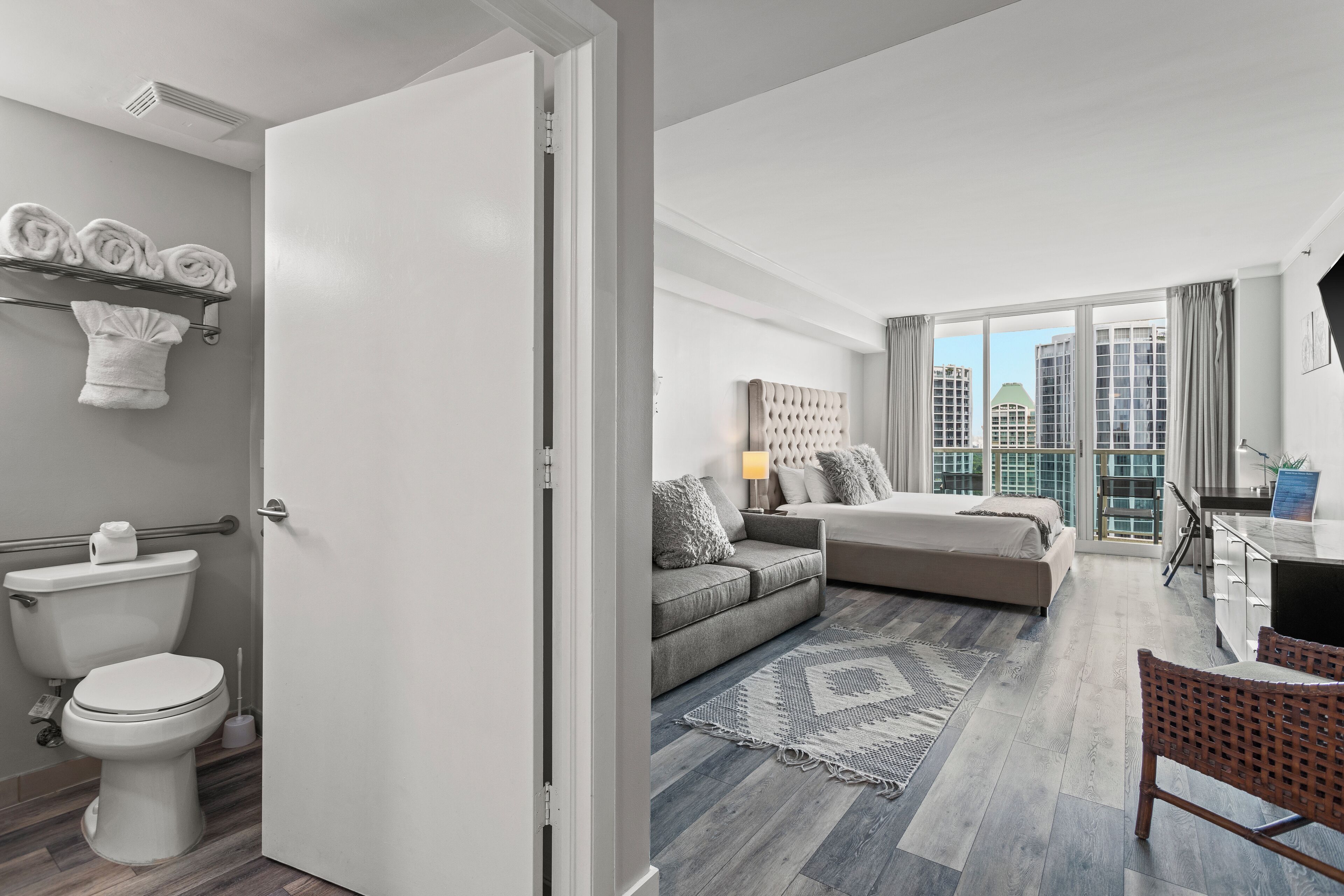 A modern apartment with a bathroom, a living area with a sofa and a dining table, and a view of the city through large windows.