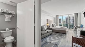 A modern apartment with a bathroom, a living area with a sofa and a dining table, and a view of the city through large windows.