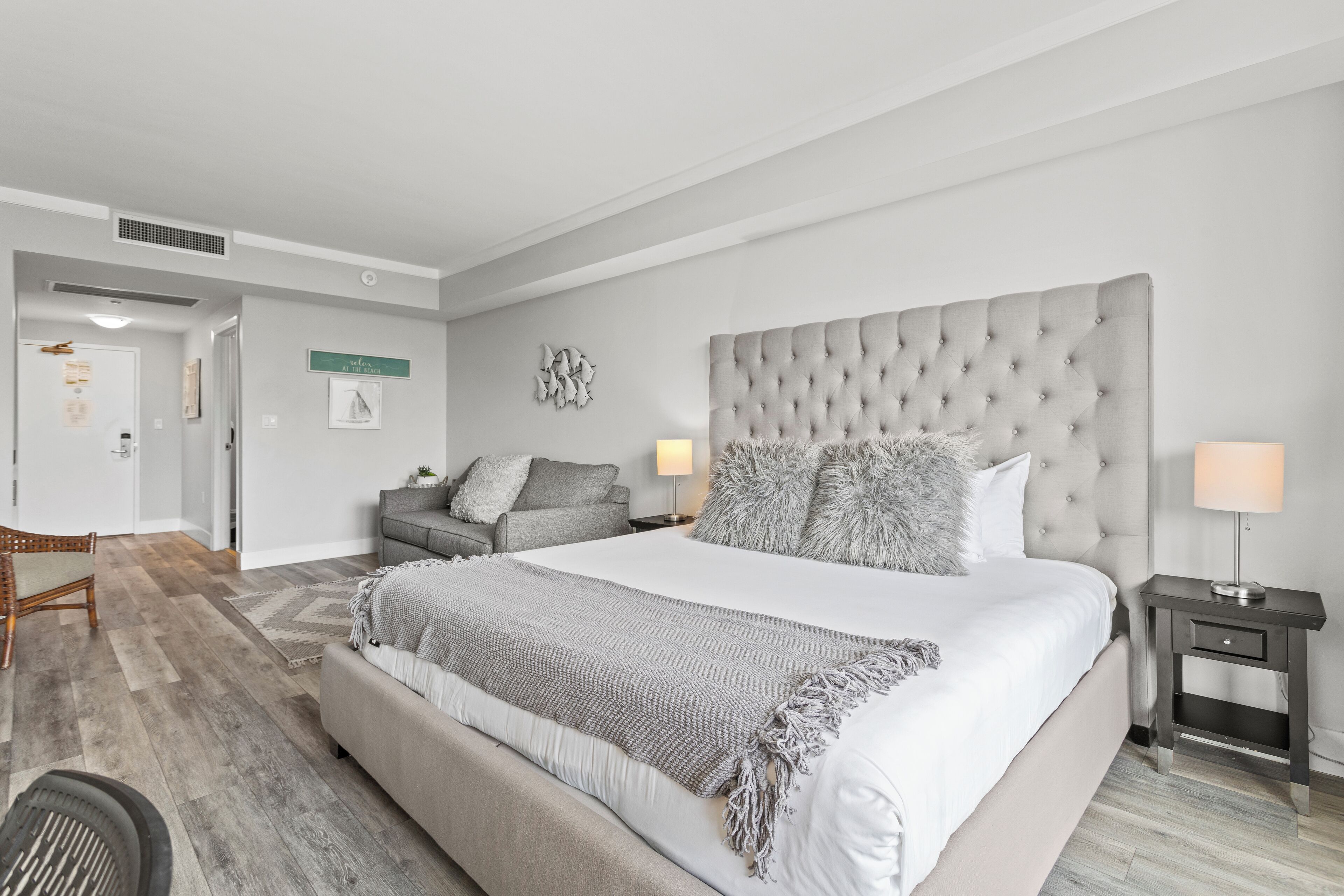 A modern hotel room with a large bed, a grey sofa, a small table, and a wall-mounted artwork.