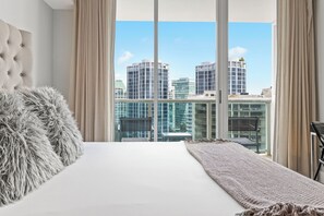A modern hotel room with a large bed, fluffy pillows, a view of tall buildings, and a balcony with curtains.