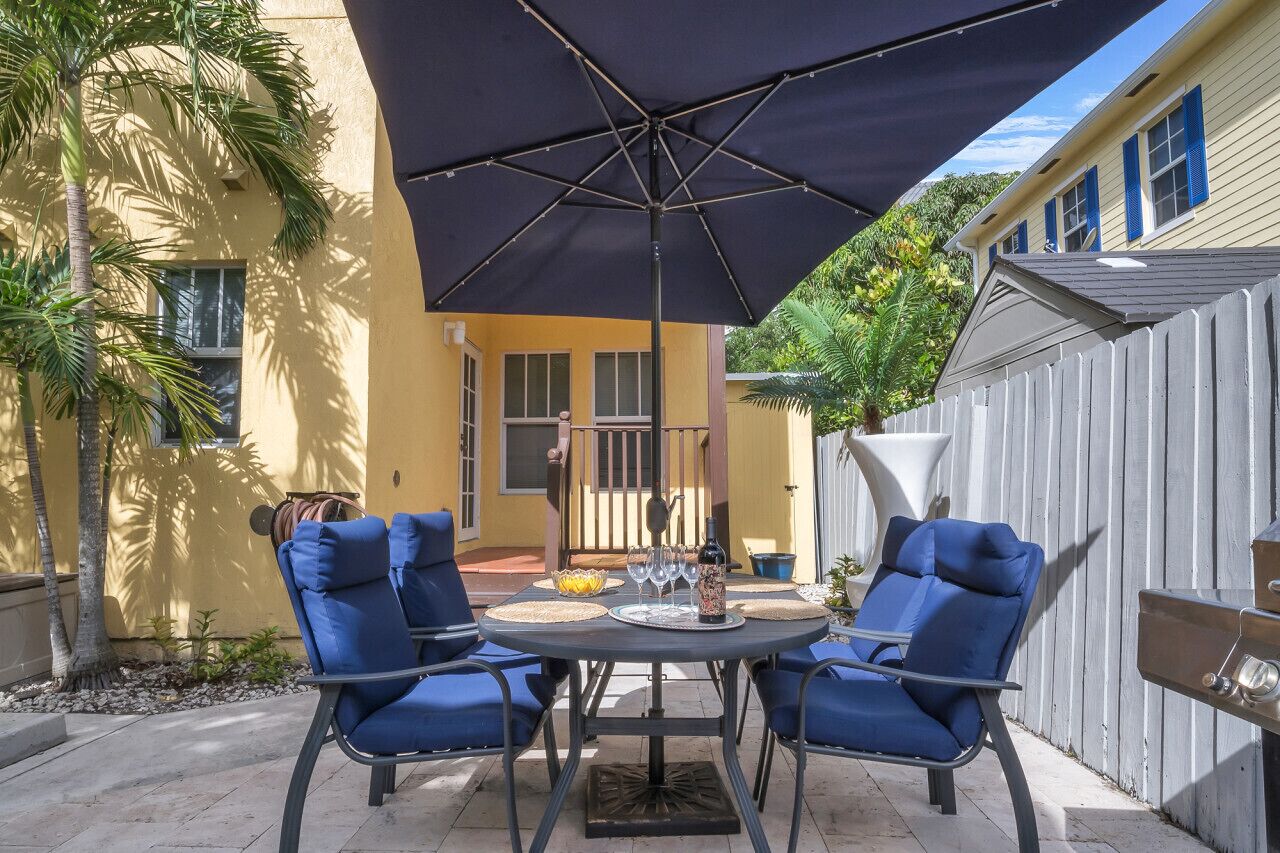 An outdoor patio with a round table, two blue chairs, a large umbrella, and a grill.