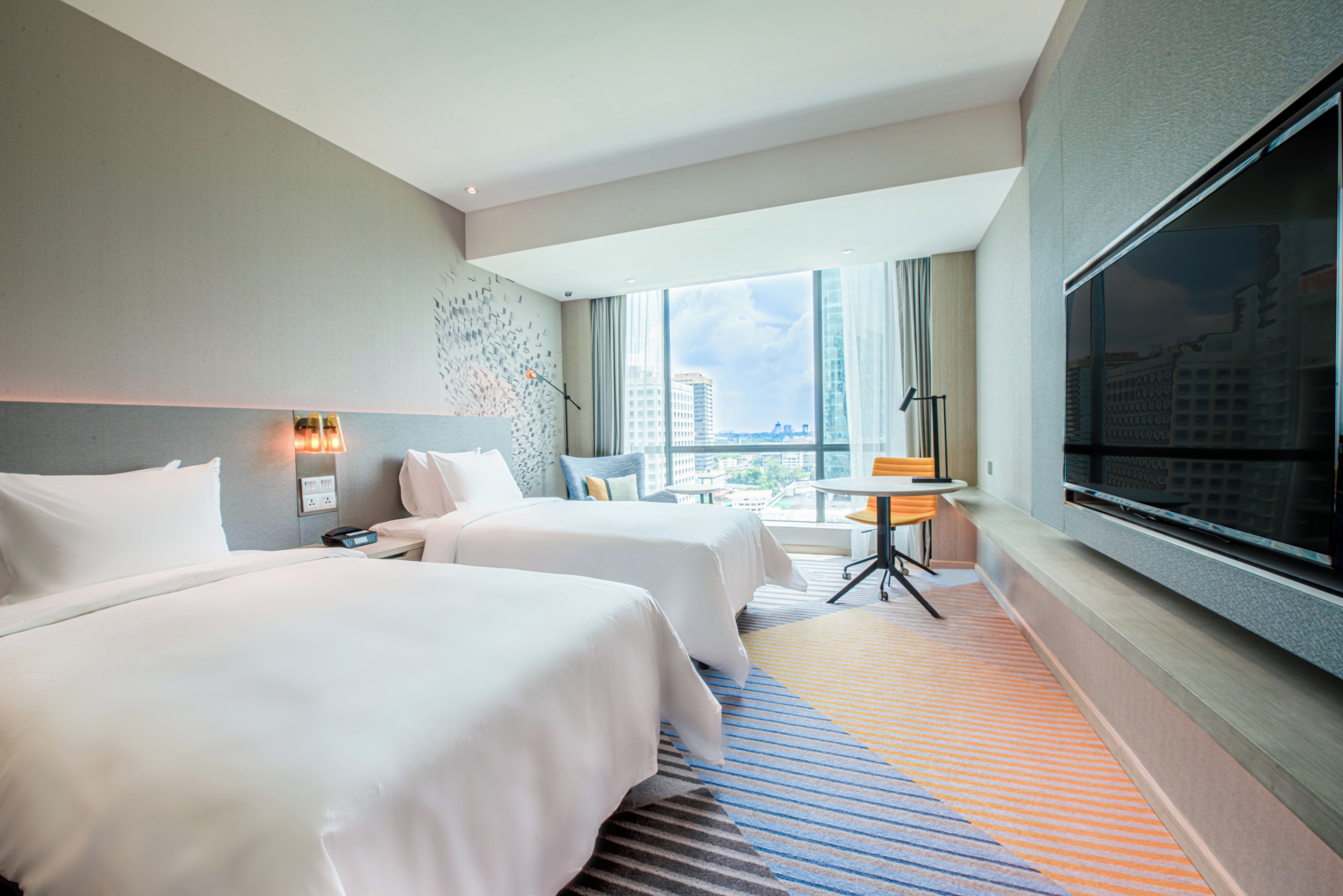 A hotel room with two beds, a television, a desk, and a view of the city.