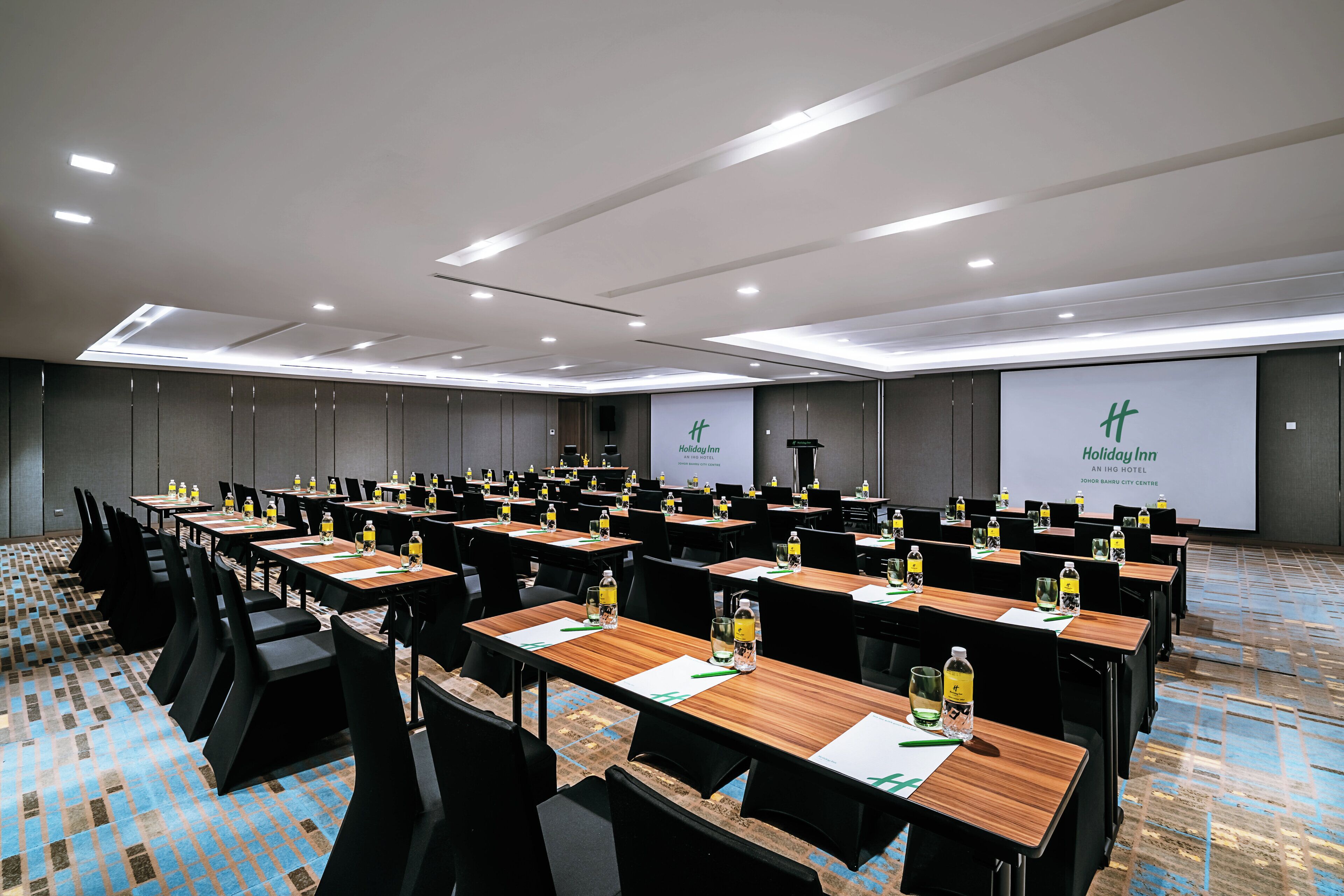 A conference room with multiple tables and chairs, equipped with water bottles and nameplates, and a projection screen displaying the hotel name.