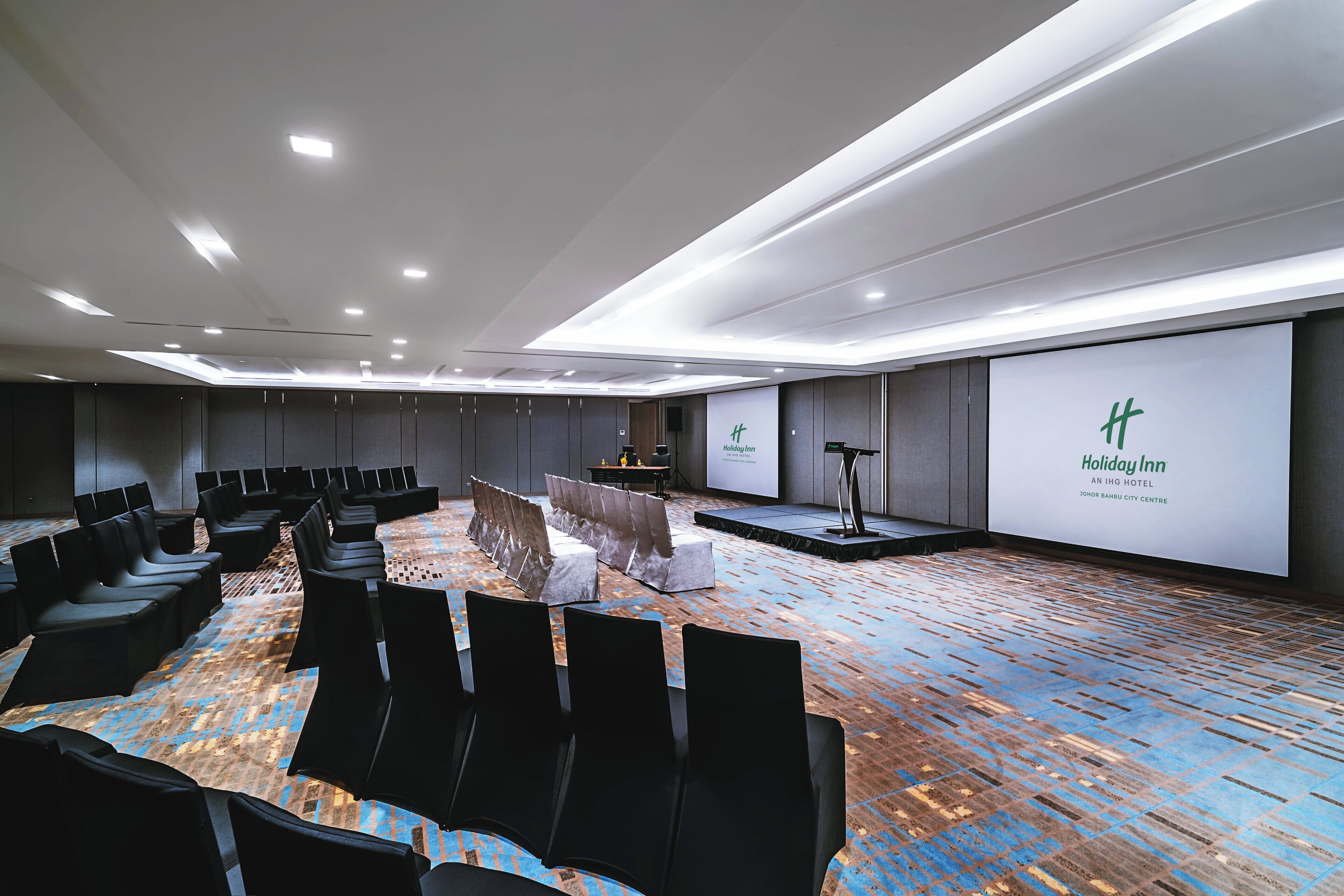 A conference room with rows of chairs, a stage, and a large screen displaying the Holiday Inn logo.