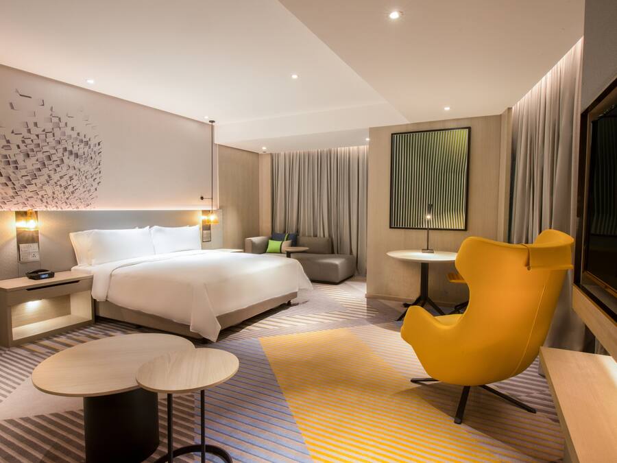 Une chambre d’hôtel moderne avec un grand lit, un canapé, un fauteuil jaune, une petite table et une grande fenêtre.