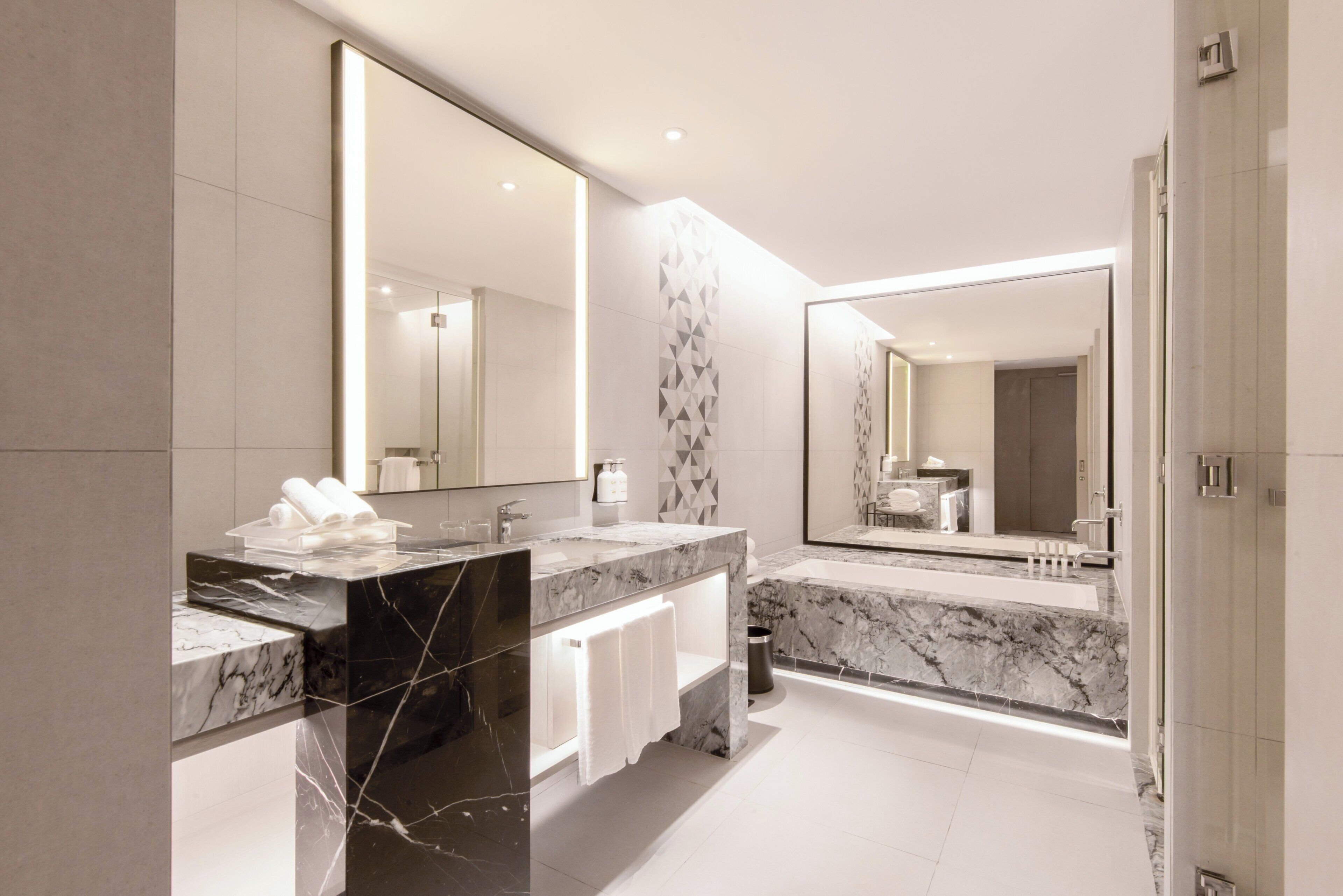 A modern bathroom with a large mirror, two sinks, marble countertops, and a bathtub.