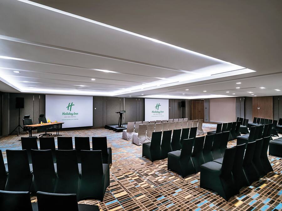 Een congreszaal met rijen zwarte stoelen, een podium en twee grote schermen waarop het Holiday Inn logo wordt getoond.