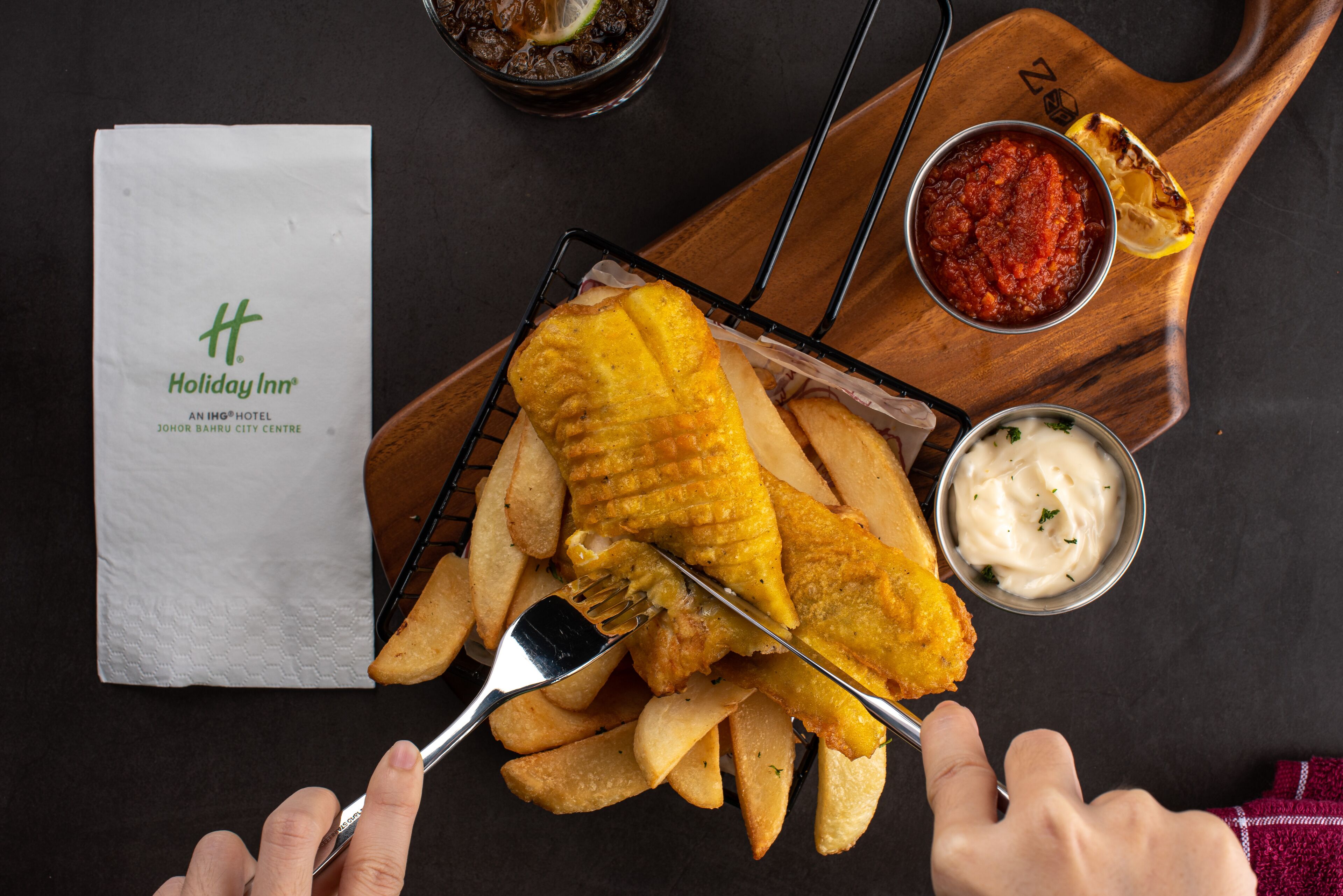 A tray of fish and chips with a side of tartar sauce and a small bowl of ketchup, accompanied by a napkin and a glass of a dark beverage.