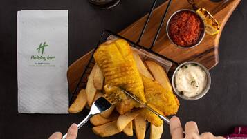 A tray of fish and chips with a side of tartar sauce and a small bowl of ketchup, accompanied by a napkin and a glass of a dark beverage.