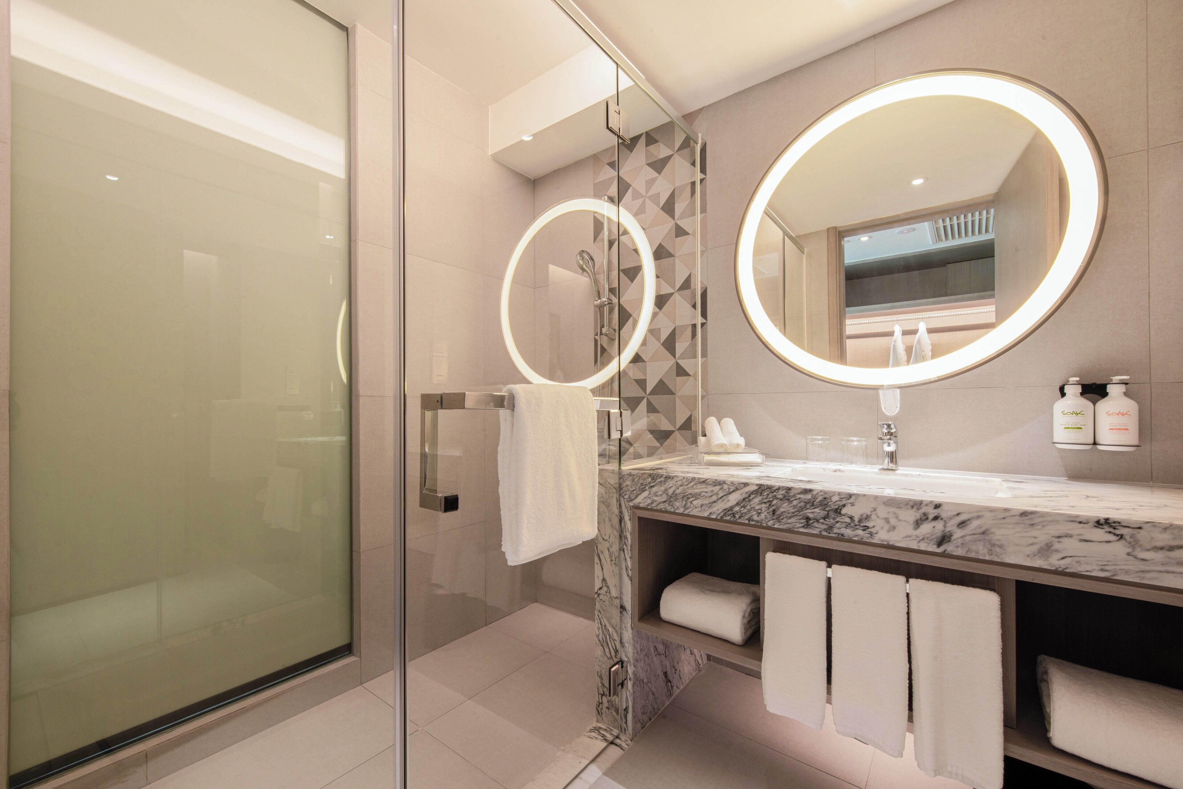 A modern bathroom with a glass shower enclosure, marble countertop, dual round mirrors, and a geometric tile backsplash.