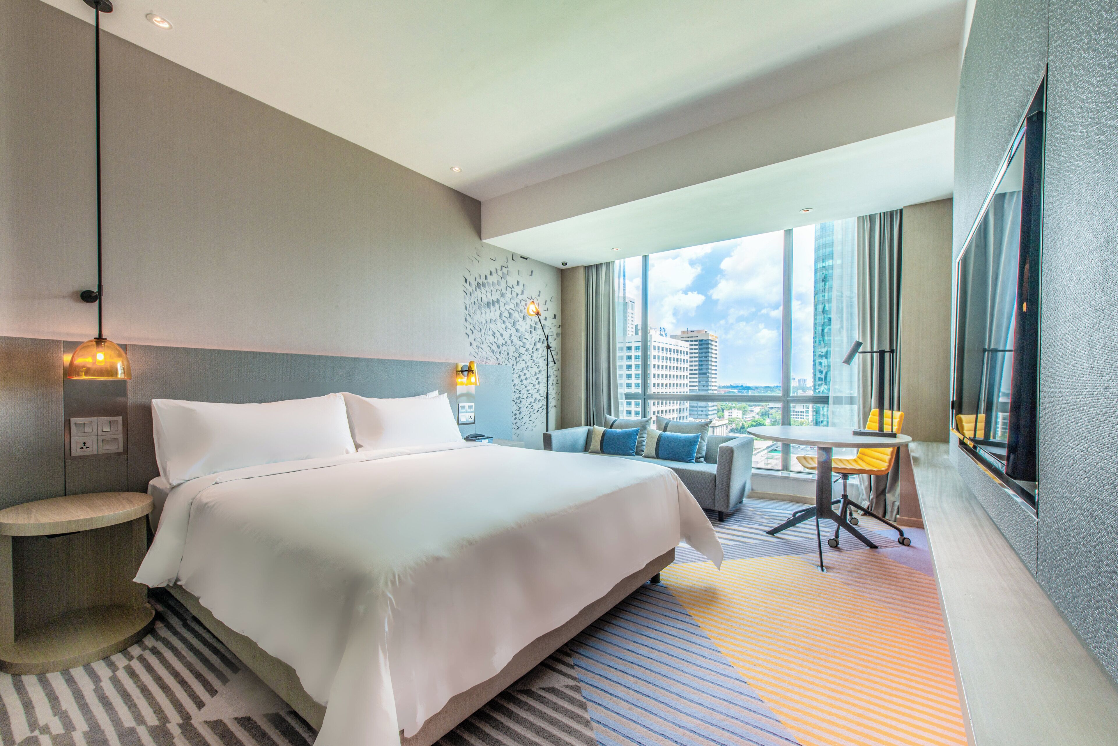 Habitación de hotel con una cama grande, zona de estar con una mesa y vistas a la ciudad a través del ventanal.