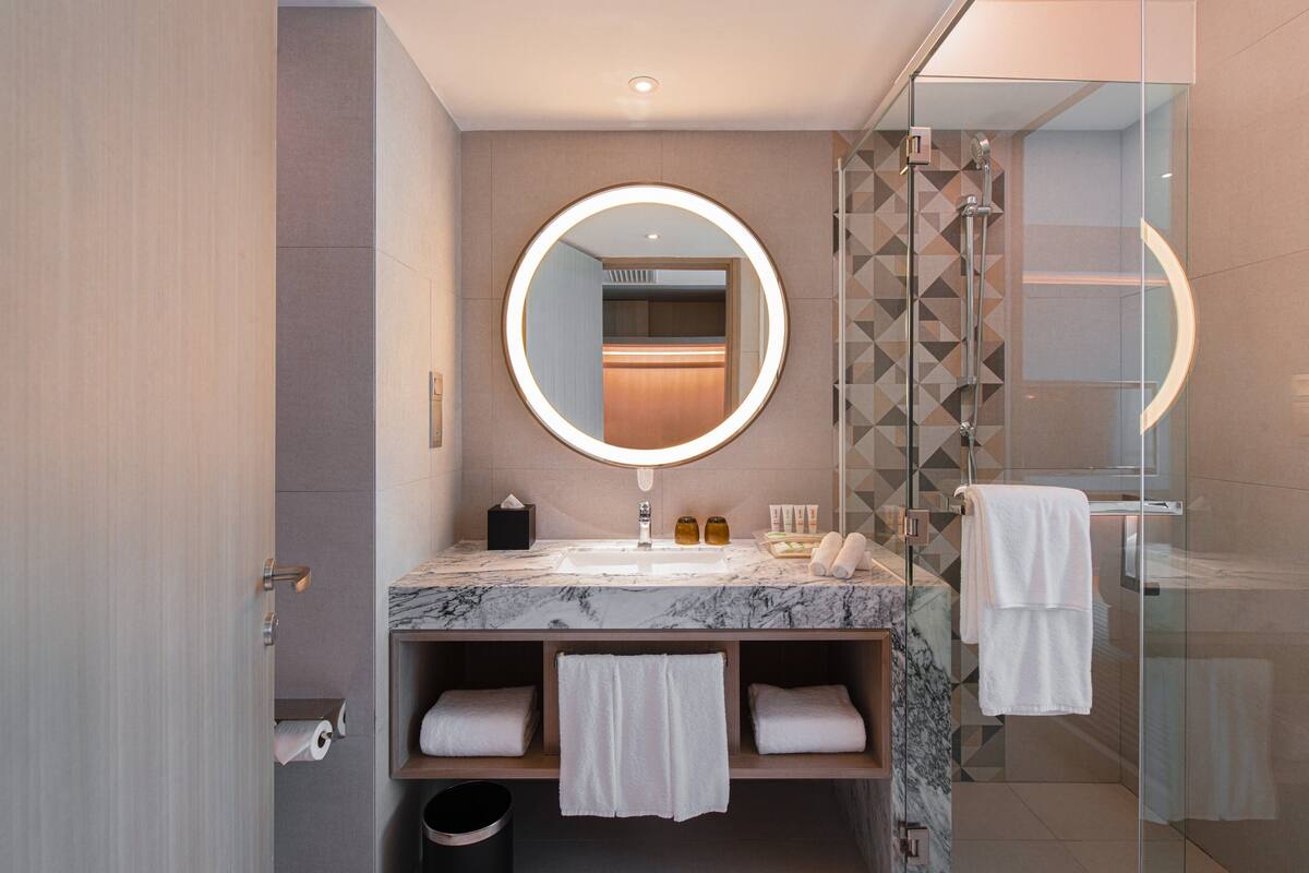A modern bathroom with a marble sink, a round mirror with a light, and a glass shower enclosure.