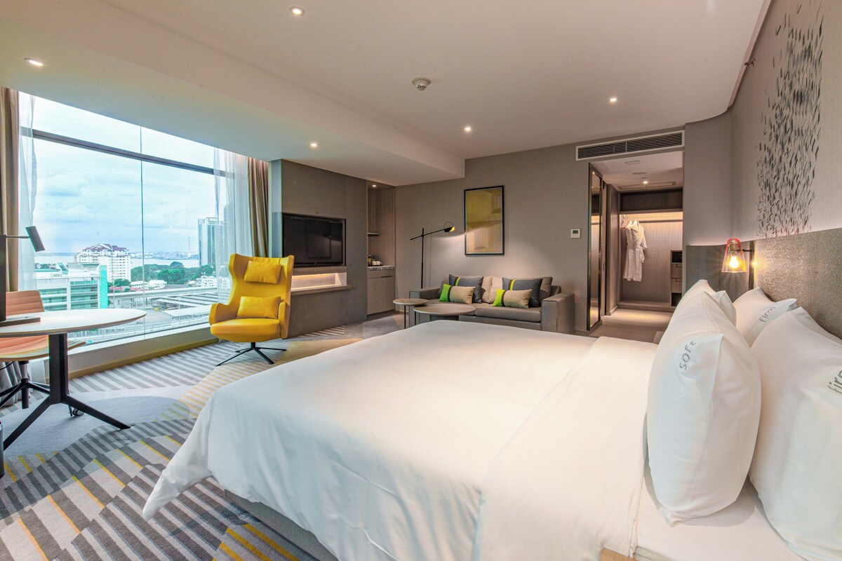A hotel room with a large bed, a sofa, a yellow armchair, a TV, and a view of the city.