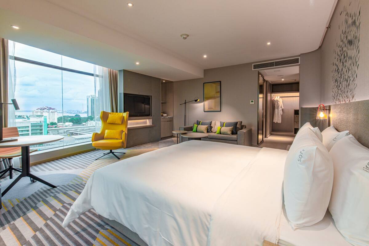 A modern hotel room with a large bed, a yellow armchair, a sofa, a small table, and a TV.