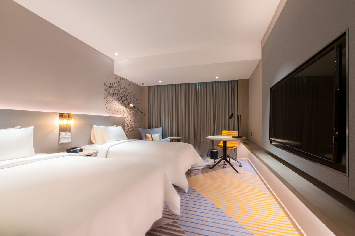 A modern hotel room with a large bed, a small table, a chair, a TV, and a large window.