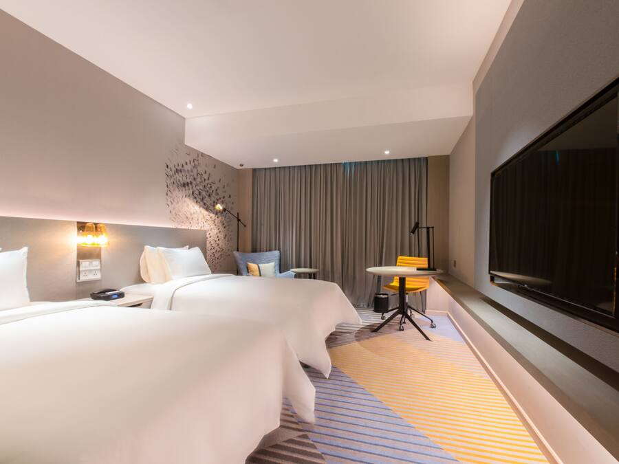 A modern hotel room with a large bed, a small table, a chair, a TV, and a large window.