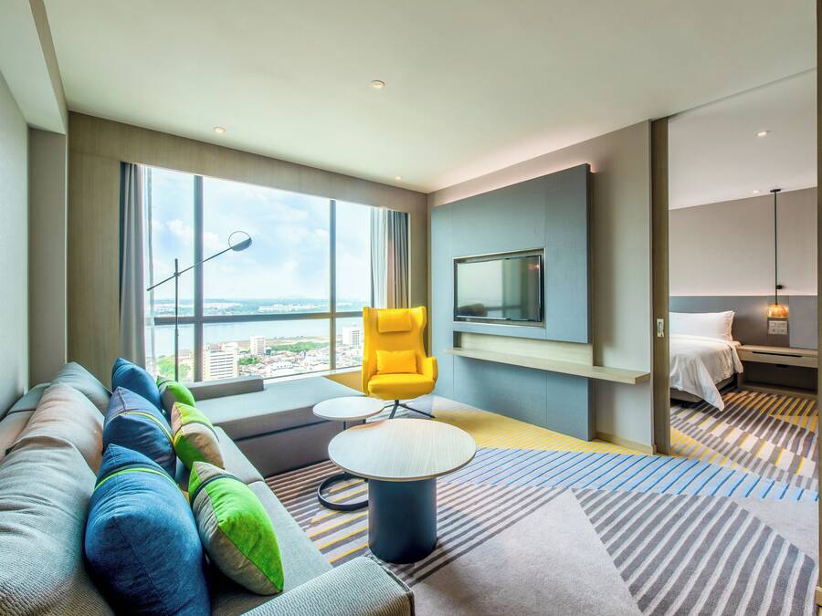 Una habitación de hotel moderna con un ventanal grande, un sofá con almohadones de colores, un sillón amarillo, una mesita redonda y una cama con un tapete a rayas.