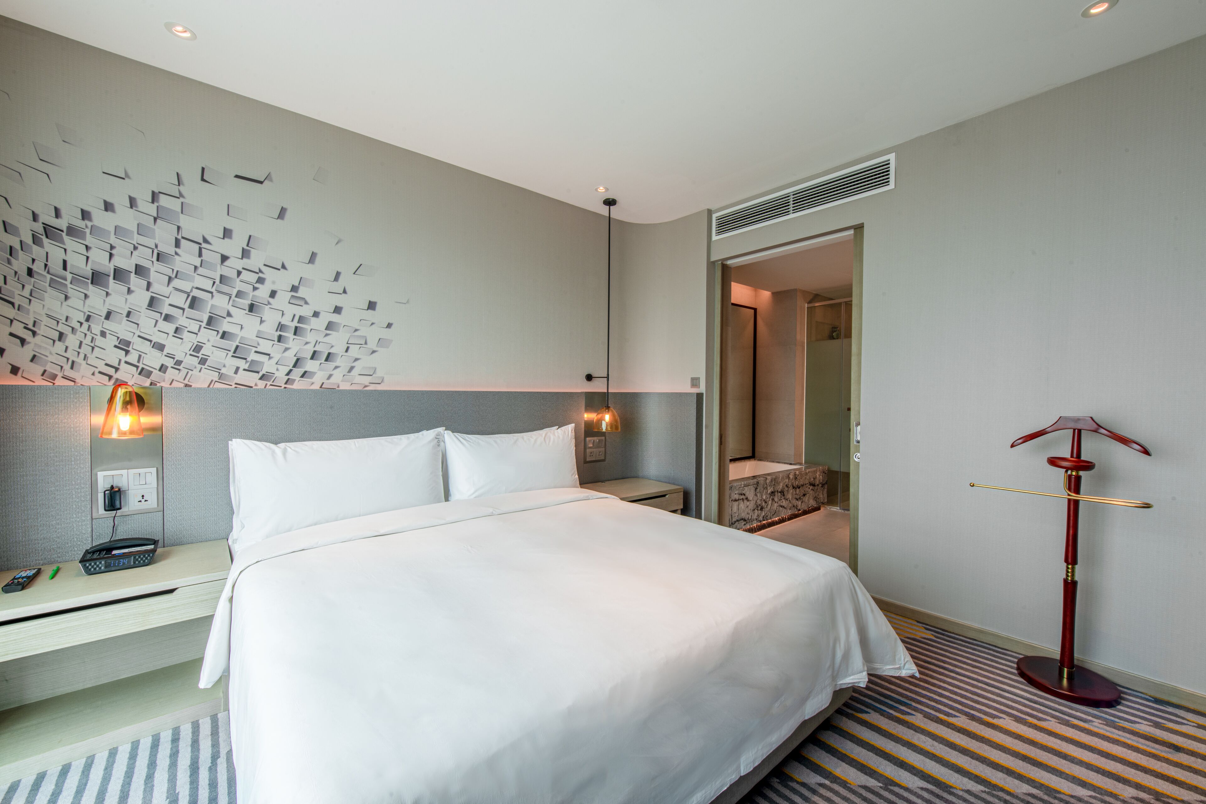 A hotel room with a neatly made bed, a bedside table with a phone and a lamp, a decorative wall panel, and a standing lamp.
