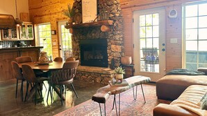 Interior acolhedor de uma cabana, com lareira de pedra, área de jantar de madeira e uma janela grande.