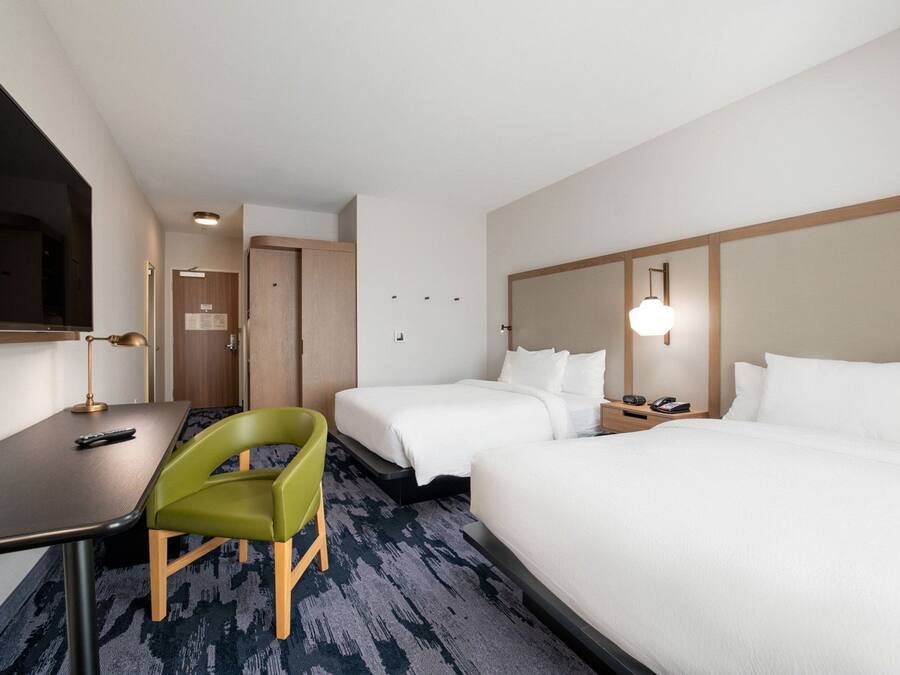 A hotel room with two beds, a desk, a chair, a TV, and a bedside table with a lamp.