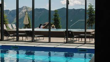 An indoor swimming pool with clear views of mountains, lounge chairs, and a small building.