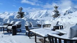 A snow-covered outdoor seating area with chairs and a trash can, set against a backdrop of snow-covered mountains and a clear sky. - Gudauri Lodge (GUDAURI)