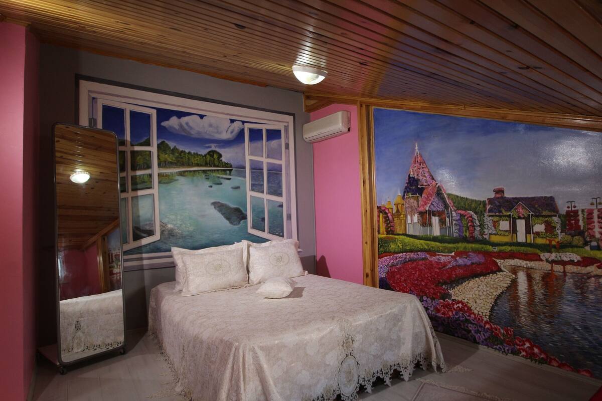 A bedroom with a bed, a mirror, a window with a view of a landscape, and a mural of a house and garden.