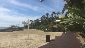 Uma quadra de voleibol de praia com rede, palmeiras e um prédio ao fundo.