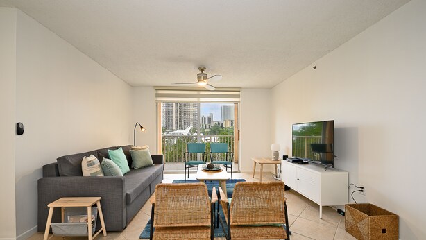 A modern living room with a grey sofa, wicker chairs, a TV, and a balcony with city views.