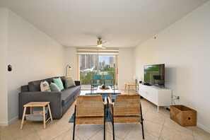 Un salón moderno con un sofá gris, sillas de mimbre, un televisor y un balcón con vistas a la ciudad.