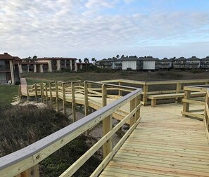 Een houten looppad dat naar een strandgedeelte leidt, met gebouwen op de achtergrond.