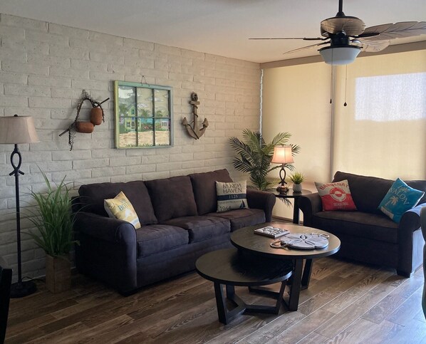 A living room with a dark brown sofa, a round coffee table, a floor lamp, a ceiling fan, and a small table with a lamp and a plant.