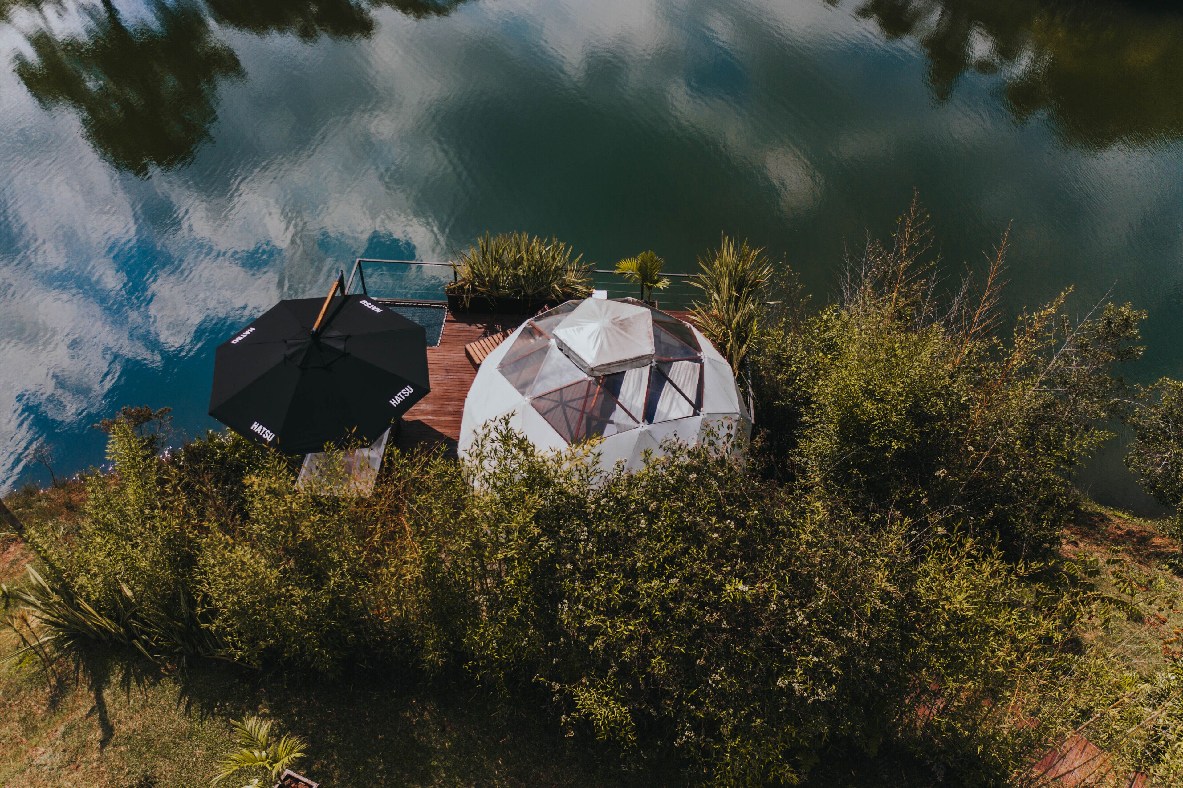 Una cabaña junto al lago con una estructura de cúpula transparente, rodeada de vegetación densa, y equipada con un paraguas negro y uno blanco.