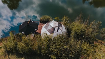 Una cabaña junto al lago con una estructura de cúpula transparente, rodeada de vegetación densa, y equipada con un paraguas negro y uno blanco.