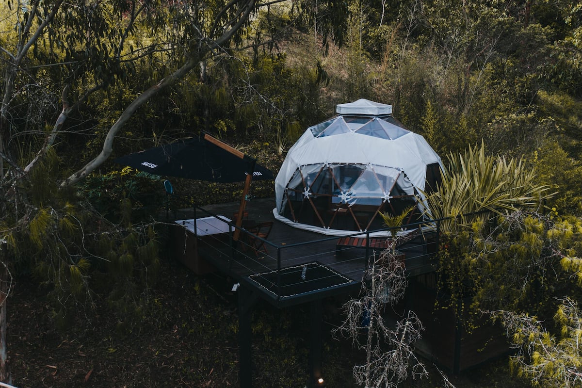A geodesic dome structure with a deck and a covered seating area, surrounded by dense forest.