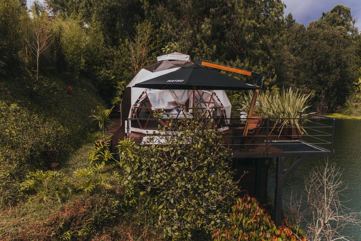 A geodesic dome structure with a transparent roof, surrounded by dense vegetation and a body of water.