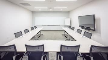 Uma sala de conferências com uma mesa comprida, cadeiras pretas, um quadro branco e uma TV na parede.