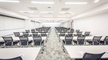 Un salón de conferencias con filas de mesas y sillas, paredes blancas y piso alfombrado.