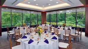 A banquet hall with round tables set for a formal event, large windows offering a view of greenery, and a modern ceiling design. - Inn at Villanova University (Wayne)