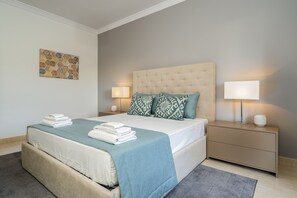A neatly made bed with a blue bedspread and two pillows, a bedside table with a lamp, and a wall-mounted artwork.
