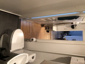 A compact bathroom with a toilet, toilet paper, and shelf, set against a backdrop of wooden paneling and a window.