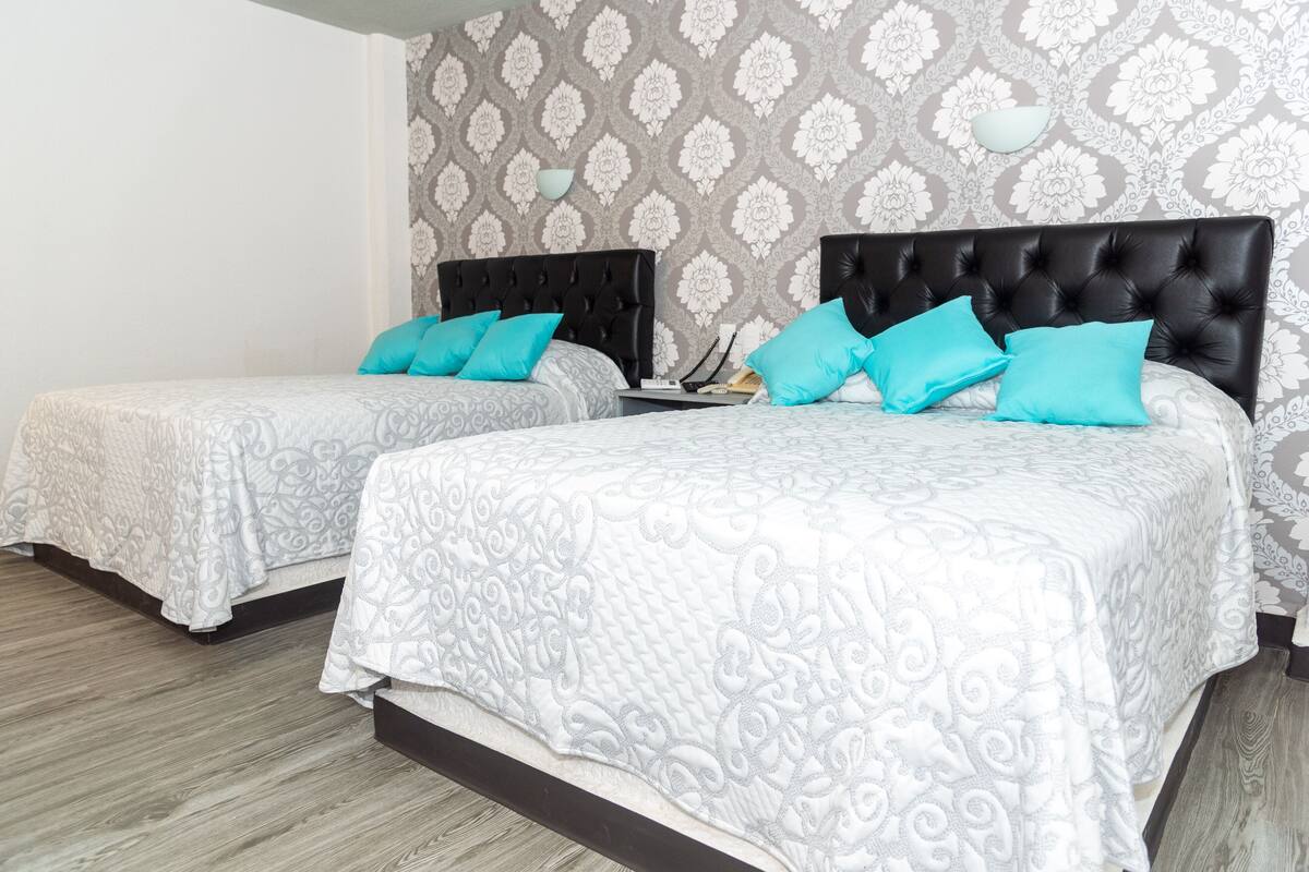 Two twin beds with matching headboards and decorative pillows, a bedside table with a lamp, and a patterned wallpaper.