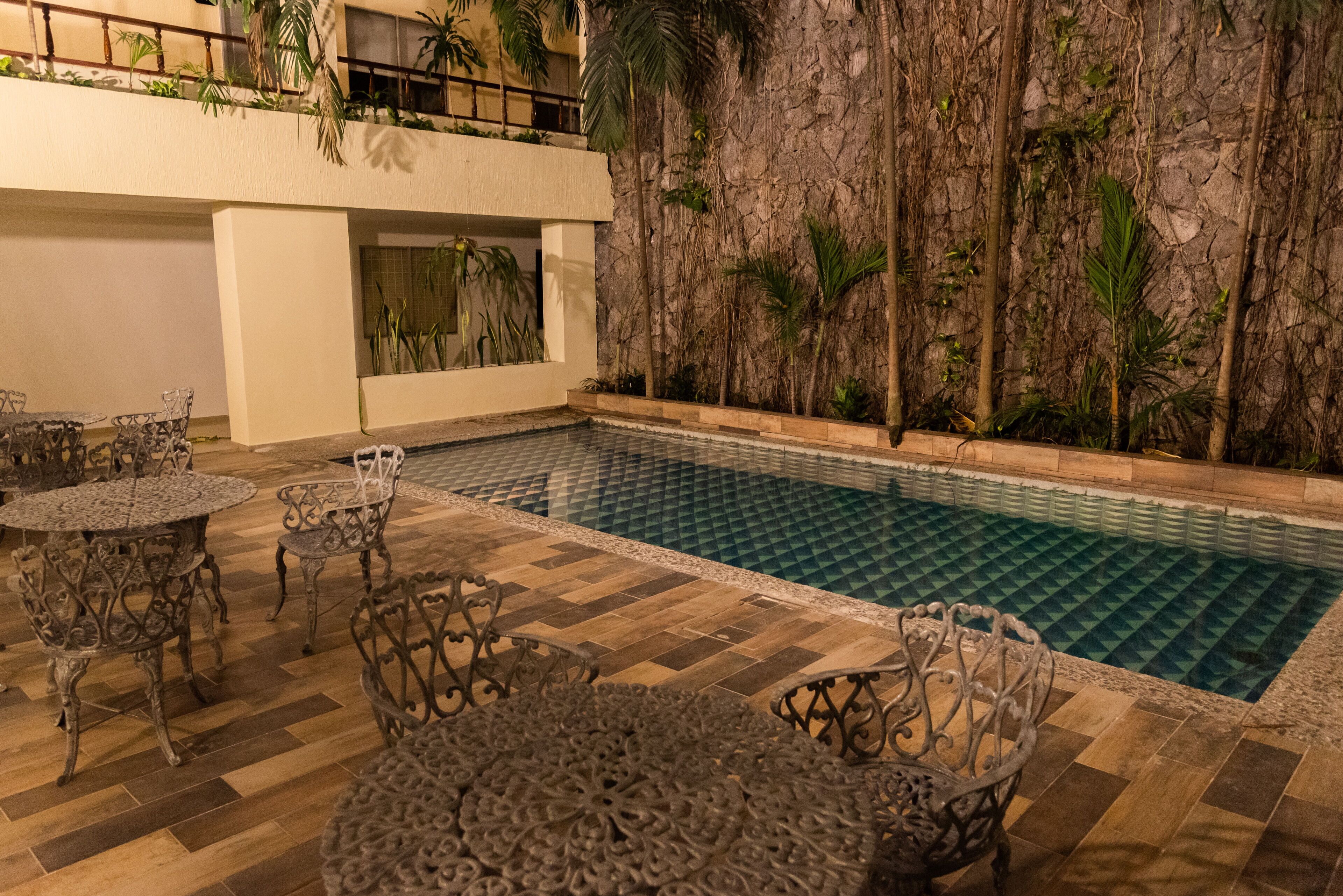 Area piscina con sedie e tavoli in metallo decorato, muro in pietra con piante e vista su un edificio con balconi.