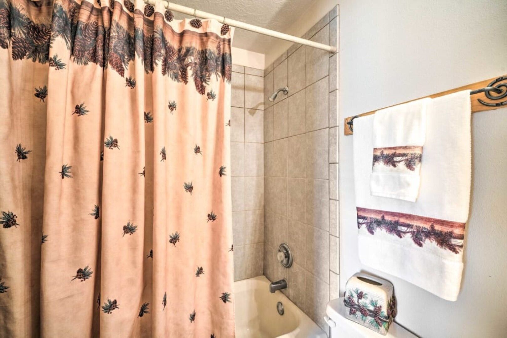 Une salle de bain équipée d’un rideau de douche orné de motifs représentant des arbres et des pommes de pin, d’une baignoire et d’un porte-serviettes avec une serviette.